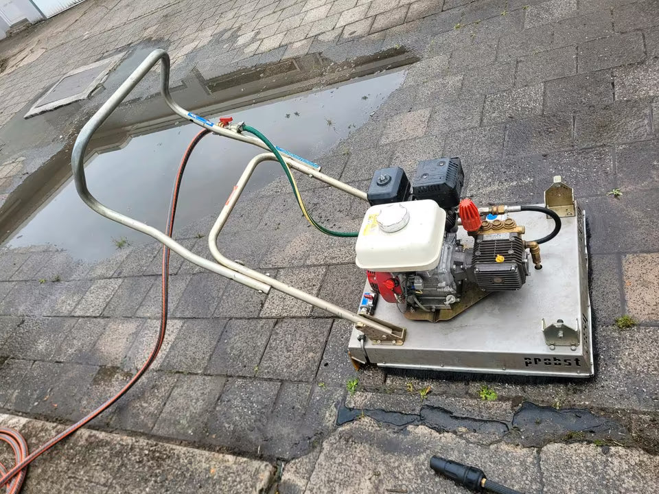 Ein weiß-silbernes Pflasterreinigungsgerät, welches ähnlich wie ein Rasenmäher geschoben wird.