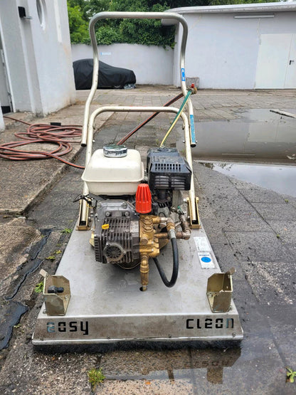 Ein weiß-silbernes Pflasterreinigungsgerät, welches ähnlich wie ein Rasenmäher geschoben wird. Man sieht hier den oben aufliegenden Motor, sowie das Gerät von Vorne.
