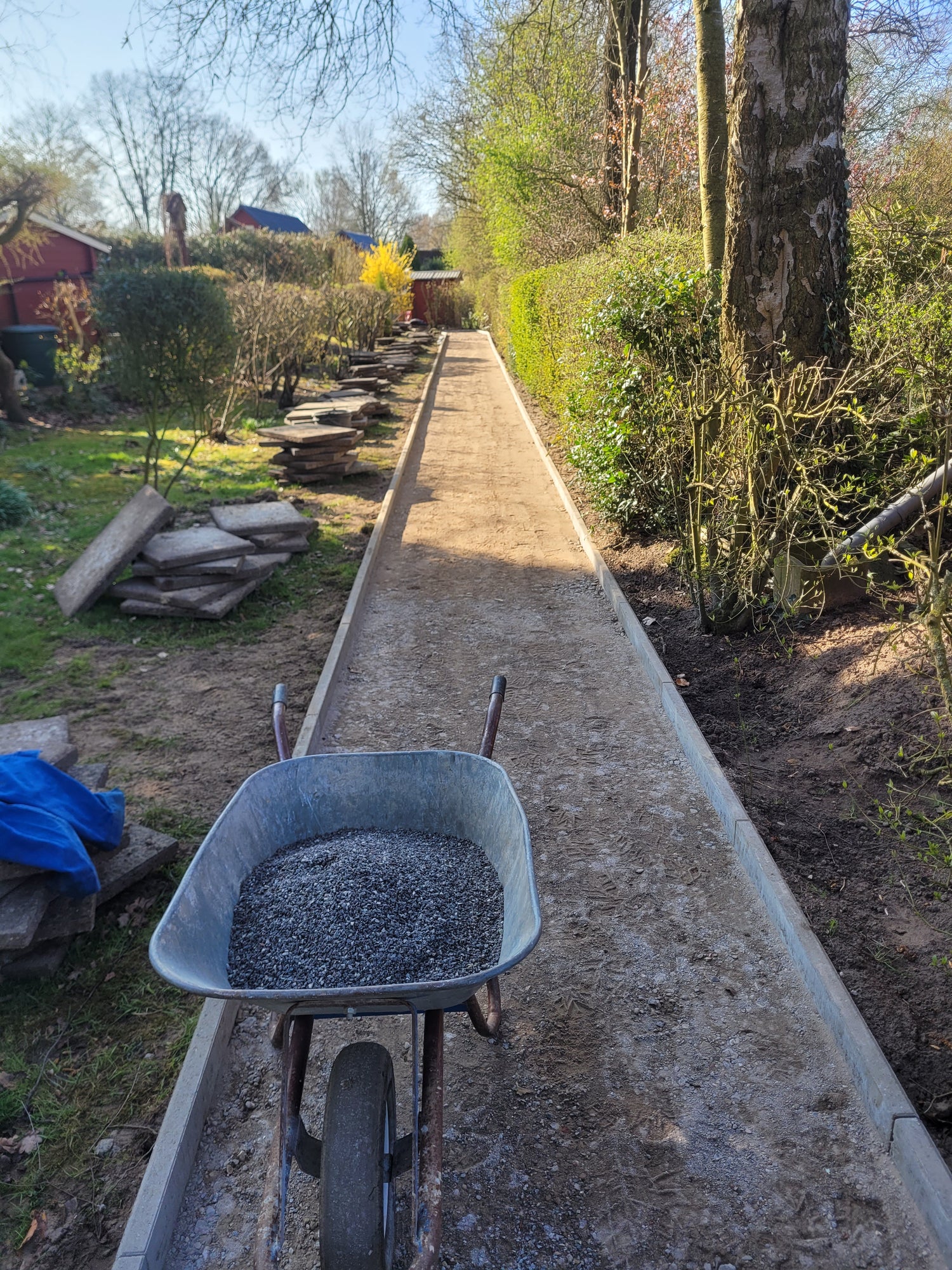 Gartenweg im Aufbau mit vorbereiteter Tragschicht, Einfassung und Schubkarre auf einer langen schmalen Wegefläche
